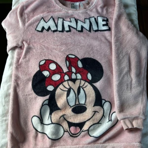 Rosa fluffig Minnie Mouse tröja H&M - Supermjuk och fluffig rosa tröja från H&M med stort tryck av Minnie Mouse och texten 'MINNIE' framtill. Tröjan har långa ärmar och är gjord i 100% polyester. Perfekt för dig vill ha något riktigt mysigt och mjukt!