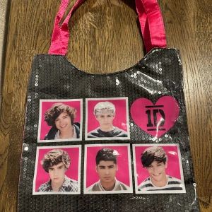 One Direction shoppingväska med paljetter - Cool shoppingväska med svart framsida täckt av glittriga paljetter, rosa baksida och handtag i satin. Framsidan har bilder på One Direction-medlemmarna och ett stort rosa hjärta med bandets logga. Perfekt för dig som vill visa din fandom.