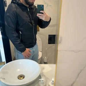 Svart vindjacka från BOSS med mönster - Snygg svart vindjacka från BOSS med diskret rutigt mönster och dragkedja framtill. Jackan har huva med snörning, två dragkedjefickor och justerbar nederkant. Perfekt för dig som vill ha en stilren jacka med coola detaljer.