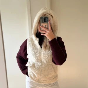 Vit fleeceväst med huva och dragkedja - Mysig vit fleeceväst med stor huva och praktisk dragkedja framtill. Västen har två fickor och är fluffig med en cool, avslappnad look. Perfekt att slänga över en hoodie eller tröja för extra värme. Passformen är normal och västen är enkel att styla.