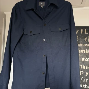 Mörkblå overshirt från H&M - Stilren mörkblå overshirt från H&M med klassisk krage, två bröstfickor med lock och knappar framtill. Skjortan har lång ärm och rak passform, perfekt att bära öppen eller stängd. Materialet känns mjukt och lätt, vilket gör den enkel att styla.