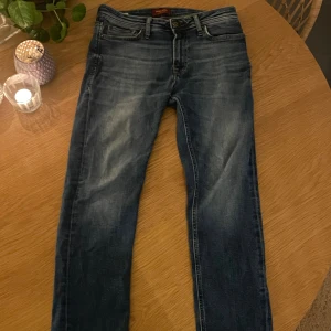 Blå jeans från Jack & Jones - Snygga blå jeans från Jack & Jones med klassisk femficksdesign och diskret slitning på framsidan. Jeansen har normal passform och raka ben, perfekt för en avslappnad stil. Tillverkade i bomull med en autentisk denimkänsla och detaljerade sömmar.