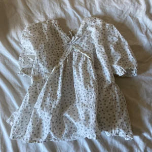 Vit blommig blus från Kappahl - Supersöt vit blus från Kappahl med små blommiga mönster i svart och beige. Klänningen har puffiga korta ärmar, knytning framtill och volangkant. Perfekt för sommarens varma dagar och har en luftig passform i mjuk bomull. 💗