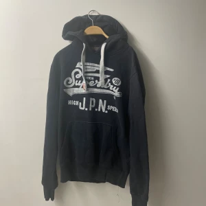 Svart Superdry hoodie med tryck - Svart hoodie från Superdry med stort vitt tryck på bröstet och grå snören i huvan. Mjuk insida i fleece och klassisk känguruficka framtill. Perfekt för dig som gillar streetwear och vill ha något bekvämt och snyggt.