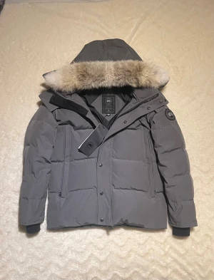 Grå Canada Goose jacka  - Säljer en grå  Canada Goose  Wyndham med avtagbar huva och pälskant. Jackan har svart logotyp på ärmen, dragkedja och knappar framtill samt ribbade muddar. Perfekt för kalla vinterdagar och riktigt snygg passform.