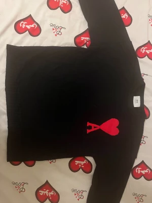 Svart stickad tröja Ami Paris med rött hjärta - Svart stickad tröja från Ami Alexandre Mattiussi med ett stort rött hjärta och bokstaven A broderat på bröstet. Tröjan har rund hals, ribbade muddar och lång ärm. Perfekt för dig som gillar stilrena detaljer med en twist. Storlek M.