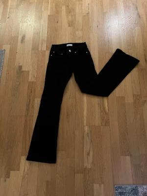 Svarta bootcut jeans med fickdetaljer - Snygga svarta jeans från Gina Tricot med bootcut perfect jeans passform. Baksidan har fickor med lock och silverfärgade knappar. Jeansen är low waist och är tillverkade i ett mjukt jeansmaterial som sitter skönt hela dagen.