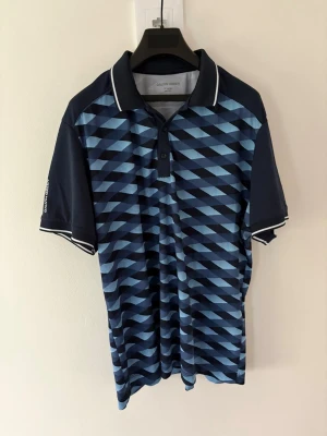 Blå mönstrad golfpiké Galvin Green - Snygg golfpiké från Galvin Green i marinblått med geometriskt mönster i olika blå nyanser. Tröjan har krage med vit kant, korta ärmar och knappar framtill. Perfekt för dig som gillar sportig och modern stil. Storlek: XL passar L. Nypris: 999kr