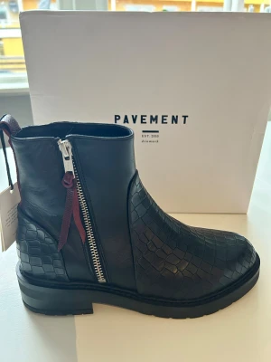 Svarta boots från Pavement med dragkedja - Snygga svarta boots från Pavement i skinn med coolt ormskinnsmönster och silverfärgad dragkedja på sidan. Skorna har rund tå, platt sula och en mörkröd detalj vid dragkedjan. 