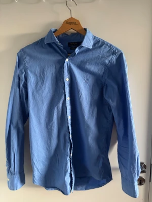Blå skjorta från Polo Ralph Lauren - Säljer en stilren blå skjorta från Polo Ralph Lauren i slim fit. Skjortan är tillverkad i tunn bomullstwill och har klassisk krage, vita knappar och långa ärmar. Perfekt för dig som gillar en clean och tidlös look.