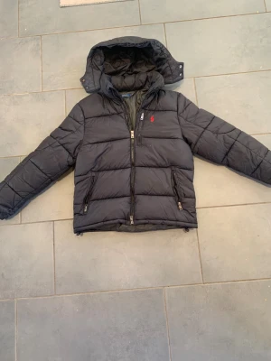 Svart dunjacka Polo Ralph Lauren - Svart dunjacka från Polo Ralph Lauren med huva och klassisk röd logga på bröstet. Jackan har quiltad design, dragkedjor på fickorna och framtill, samt en normal passform. Perfekt för kalla dagar och riktigt snygg till streetwear. Påsen och taggarna kommer inte med köpet