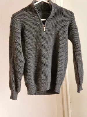 Mörkgrå stickad tröja med half zip - Säljer en mörkgrå stickad tröja med ribbad struktur och half zip i halsen. Tröjan har långärmad design och en hög krage som ger extra värme. Perfekt för dig som gillar en enkel men snygg look under kyliga dagar.