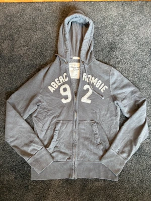 Grå zip-hoodie Abercrombie & Fitch  - Snygg grå hoodie från Abercrombie & Fitch i storlek S. Tröjan har broderade vita detaljer med siffran 92 och märkesnamnet på bröstet, huva med snörning, två fickor fram och dragkedja. Perfekt för chill dagar och streetstyle.