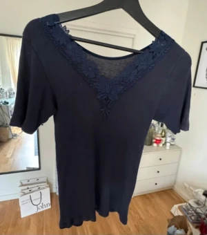 Mörkblå blus med spetsdetaljer - Säljer en mörkblå blus med korta ärmar och v-ringad hals med snygga spetsdetaljer i blommönster. Blusen har en mjuk och lätt struktur, perfekt för dig som gillar stilrena plagg med en twist. Passformen är normal och materialet känns som bomull.