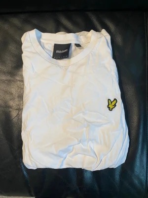 Vit t-shirt från Lyle & Scott - Vit t-shirt från Lyle & Scott med klassisk rund hals och broderad gul logga på bröstet. Tillverkad i mjuk bomull och har en enkel, stilren design som passar perfekt till jeans eller shorts. Den har dock ett litet hål på magen som knappt syns.