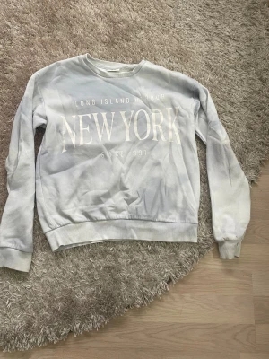 Ljusgrå sweatshirt New York Kappahl - Snygg ljusblå sweatshirt från Kappahl med stort vitt tryck 'NEW YORK' och texten 'Long Island Harbor Since 1991' på framsidan. Tröjan har rund hals, ribbade muddar. Perfekt för dig som gillar streetstyle och chill vibes.
