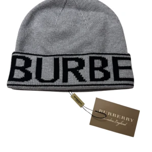 Grå stickad Burberry mössa - Snygg grå stickad mössa från Burberry med bred svart logotyp runt kanten. Klassisk form med uppvikt kant och tydlig Burberry-tag. Perfekt för dig som vill ha en stilren och lyxig accessoar med street-vibe.