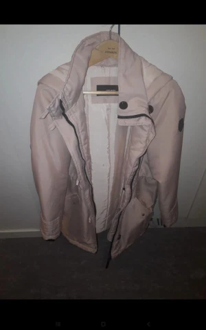 Ljusrosa parkasjacka med fuskpäls - Snygg ljusrosa parkasjacka från Vero Moda med avtagbar fuskpäls till huvan. Jackan har dragkedja, stora knappar och praktiska fickor. Perfekt för kyligare dagar och har en klassisk, lite längre modell.