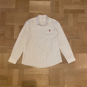 Beige randig skjorta från Ami - Snygg beige skjorta från Ami Alexandre Mattiussi i storlek L med svarta ränder och röd broderad logga med hjärta och A på bröstet. Skjortan har klassisk krage, långa ärmar och knappar framtill. Perfekt för dig som gillar stilrena detaljer och fransk design. Använd endast 4 gånger vid speciella tillfällen. Tvättad efteråt.