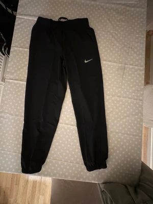 Svarta Nike Dri-FIT träningsbyxor - Svarta träningsbyxor från Nike med Dri-FIT-teknologi, elastisk midja med snörning och dragkedjor vid bensluten. Byxorna har en slim passform och klassisk Nike Swoosh på låret. Perfekta för träning eller chill.