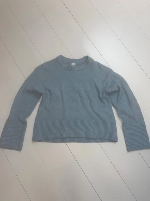 Ljusblå stickad tröja med breda muddar - Säljer en ljusblå stickad tröja med rund hals och breda ribbade muddar vid ärmslut och nederkant. Tröjan har en loose passform och är långärmad, perfekt för att hålla sig varm och snygg under kyliga dagar.