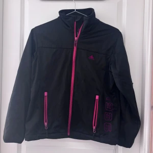 Svart softshelljacka från Likkia 158/164 - Svart softshelljacka från Likkia med rosa dragkedjor och detaljer. Jackan har hög krage, två fickor med dragkedja och tryckt siffra 1996 på ena sidan. Perfekt för dig som gillar sportig och stilren look.