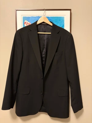 Svart kavaj Oscar Jacobson Edmund 50 - Klassisk svart kavaj från Oscar Jacobson, modell Edmund Blazer. Tillverkad i exklusivt ulltyg från Vitale Barberis Canonico, med två knappar framtill, fickor med lock och innerfoder med logga. Snygg passform och tidlös design, perfekt för en stilren look.