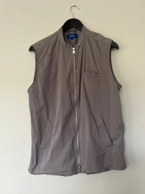Grå väst från Eton med dragkedja - Stilren grå väst från Eton med rak passform och hög krage. Västen har en praktisk dragkedja framtill, två snedställda fickor och en bröstficka. Tillverkad i lätt polyester som ger en clean look. 62 % bomull, 38 % polyester. 