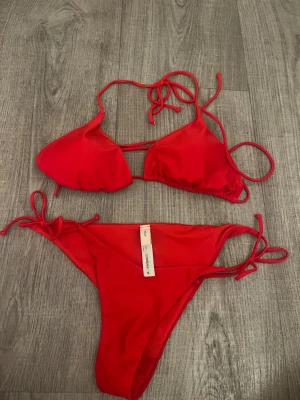 Röd bikini med knyt från Nelly - Snygg röd bikini från Nelly i storlek S. Klassisk trekantsmodell med knyt både i ryggen och vid höften. Mjukt syntetmaterial som sitter skönt och ger en clean look. Perfekt för stranden eller poolen, enkel att justera med snörning.