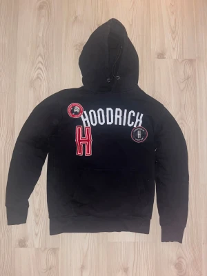Hoodrich hoodie  - Knappt använd svart hoodie från Hoodrich med stor vit logotyp och röda detaljer framtill. Tröjan har huva med snörning, lång ärm och känguruficka. Märkets patchar och broderad röd H ger en cool streetkänsla. Perfekt för dig som gillar urban stil.