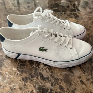 Vita sneakers från Lacoste - Snygga vita sneakers från Lacoste med klassisk krokodillogga på sidan. Skorna har snörning, rund tå och detaljer i mörkblått vid hälen. Tillverkade i skinn med vit sula och diskreta sömmar för en clean look.