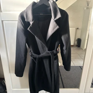 Svart kappa med bälte från Bik Bok XS - Stilren svart kappa från Bik Bok i storlek XS. Kappan har klassisk krage, långa ärmar och knyts med bälte i midjan för en snygg siluett. Perfekt för kyligare dagar och enkel att matcha med olika outfits.