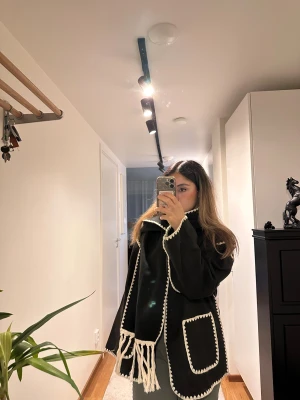 Svart overshirt med vita detaljer.Höst jacka - Svart overshirt med kontrasterande vita virkade kanter och stora fickor framtill. Jackan har en lös passform och långa ärmar. Medföljer en matchande svart scarf med vita fransar. Perfekt för dig som gillar en chill och unik stil.