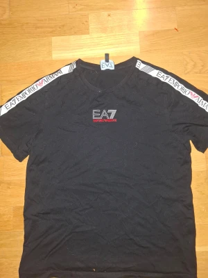 Svart EA7 Emporio Armani t-shirt - Svart t-shirt från EA7 Emporio Armani med logga broderad på bröstet och vita band med tryckt logga längs axlarna. Klassisk rund hals och korta ärmar. Materialet är mjuk bomull som känns skönt mot huden. Snygg och stilren design med sportig touch.