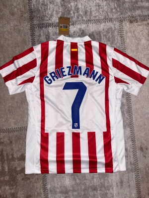 Atletico Madrid Griezmann #7 matchtröja - Säljer en officiell Atletico Madrid fotbollströja från Nike med Griezmann och nummer 7 på ryggen. Tröjan har klassiska röda och vita vertikala ränder, korta ärmar, klubbmärke på bröstet och sponsortryck. Materialet är lätt och andas, perfekt för match eller träning.