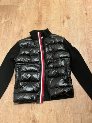 Moncler Cardigan  - Svart pufferjacka från Moncler med glansig dunfront och stickade ärmar. Jackan har två dragkedjefickor, hög krage och en snygg rödvit rand längs dragkedjan. Moncler-logga på vänster ärm. Perfekt för dig som gillar stilren och sportig look.