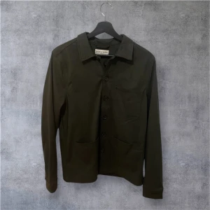 Mörkgrön overshirt från Studio Total - Snygg mörkgrön overshirt från Studio Total i storlek S. Skjortan har klassisk krage, tre framfickor och knäppning med svarta knappar. Perfekt att bära som lager över t-shirt. Materialet är mocca liknande 