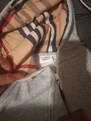 Grå Burberry hoodie med dragkedja - Snygg grå hoodie från Burberry med klassiskt rutigt foder i huvan. Tröjan har dragkedja framtill, snörning i huvan och ribbade muddar. Perfekt för dig som vill ha en stilren och lyxig look med ikonisk Burberry-detalj.