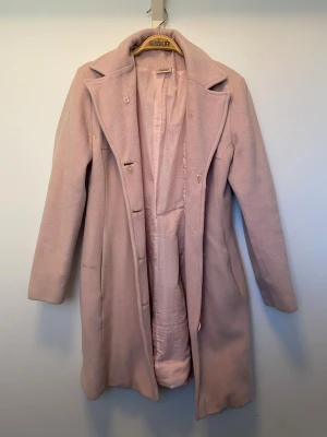Ljusrosa Kappa - Snygg ljusrosa kappa med klassisk krage och knäppning framtill. Jackan är väl använd men fortsatt i bra skick. Brukade ha en krage i fejk-päls men inte längre. Hängandes i garderoben sen en till tillbaka och det skulle vara fint att låta den få ett nytt hem! 