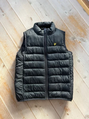 Svart dunväst från Lyle & Scott - Svart dunväst från Lyle & Scott med gul logga på bröstet. Västen har quiltad design, hög krage och dragkedja framtill. Perfekt för lager-på-lager och enkel att matcha med andra plagg.