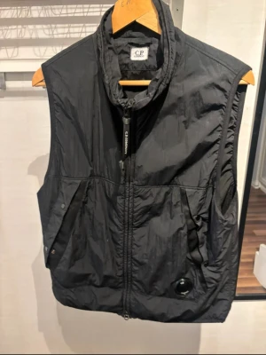 Svart Polartec väst från C.P. Company - Svart väst från C.P. Company med Polartec-material, perfekt för lager-på-lager. Väst med hög krage, dragkedja framtill och praktiska fickor. Snygg detalj med C.P. Company-logga och lins på ena fickan. Passar dig som gillar streetwear och funktion. Köpt på Johnells november 2024 Blå färgstänk på framsidan, liten prick som inte syns vid användning, även liten del på baksidan av axeln, I övrigt bra skick