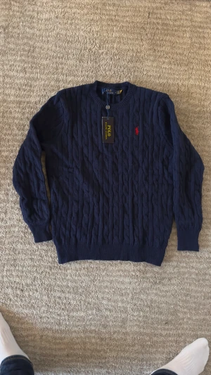 Mörkblå stickad tröja Polo Ralph Lauren - Snygg mörkblå stickad tröja från Polo Ralph Lauren med klassiskt kabelmönster och röd broderad logga på bröstet. Rund hals och ribbade muddar vid ärmslut och nederkant. Perfekt för dig som gillar stilrena och tidlösa plagg.