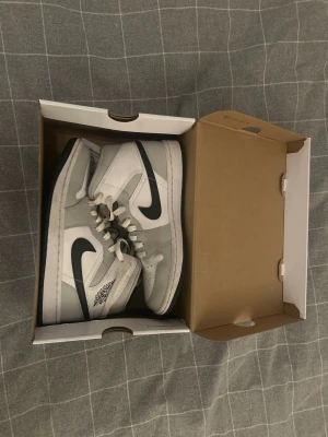 Nike Jordan 1 Mid vit/grå sneakers - Nike Jordan 1 Mid sneakers i vitt och ljusgrått med svart swoosh. Klassisk high-top siluett med snörning och rund tå. Ikonisk Air Jordan-logga på sidan. Skorna är tillverkade i skinn och syntetmaterial för en clean och stilren look.