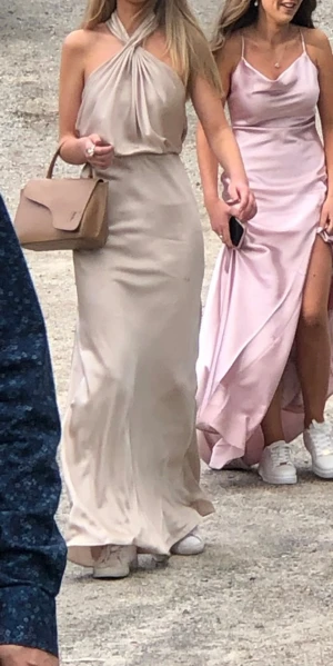 Champagnefärgad balklänning - Supersnygg långklänning i champagnefärgad satin med halterneck och draperad överdel. Klänningen har en elegant, figurnära passform och faller snyggt längs kroppen. Perfekt för student eller bal! Säljes då jag inte längre behöver den. Den har en liten fläck men de går att tvätta bort