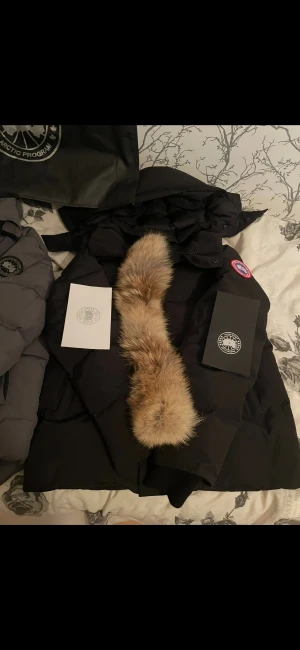  Canada Goose - Säljer en svart dunjacka från Canada Goose med ikonisk patch på ärmen och en stor fluffig päls på huvan. Jackan har dragkedja och flera praktiska fickor. Perfekt för kalla vinterdagar och har en riktigt snygg look. Wyndham parka