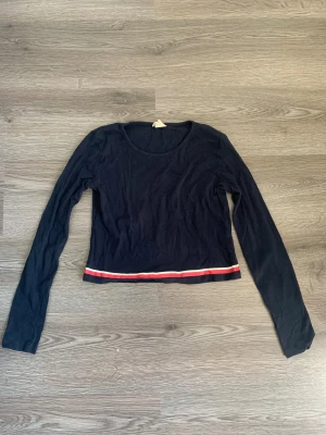 Mörkblå croppad långärmad topp - Säljer en mörkblå croppad långärmad topp i mjuk ekologisk bomull. Toppen har en rund halsringning och en snygg detalj med röd och vit rand längs nederkanten. Perfekt för dig som gillar enkel och stilren design.
