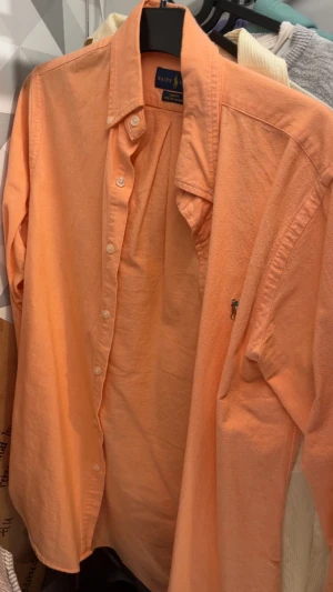 Orange skjorta från Ralph Lauren - Snygg orange skjorta från Ralph Lauren med klassisk krage och knäppning framtill. Skjortan har lång ärm och den ikoniska broderade loggan på bröstet. Perfekt för dig som vill sticka ut med färg och stil.