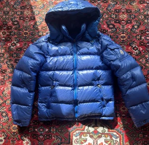  Moncler Maya. Blå med huva, strorlek 164(; - Unik Moncler-jacka i fin blå färg! Storlek 164, men passar även om du brukar ha lite större storlek. Den ljusa färgen gör att jackan är lite känslig för vatten, så det finns några små fläckar – men inget som syns direkt eller stör helheten. Jackan ser fortfarande väldigt fin ut. Hör av dig om du har frågor eller vill ha fler bilder!