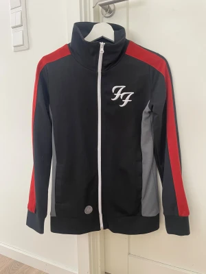 Foo Fighters svart track jacket - Svart track jacket med röda och grå detaljer från Foo Fighters. Jackan har vit dragkedja, hög krage och broderad logga på bröstet samt stort Foo Fighters-tryck på ryggen. Tillverkad i mjukt syntetmaterial, perfekt för dig som vill sticka ut med bandmerch.