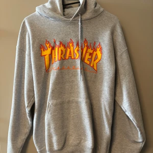 Grå Thrasher hoodie med flames - Säljer en grå hoodie från Thrasher med ikoniskt flames-tryck i gult och rött på bröstet. Tröjan har huva med snörning, stor magficka och är tillverkad i mjuk bomull och polyester. Perfekt för dig som gillar streetwear och skate-vibe. Använd enstaka gång